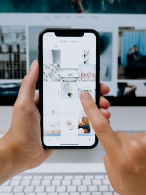 Hands holding a smartphone while browsing a social media app in front of a desktop computer.